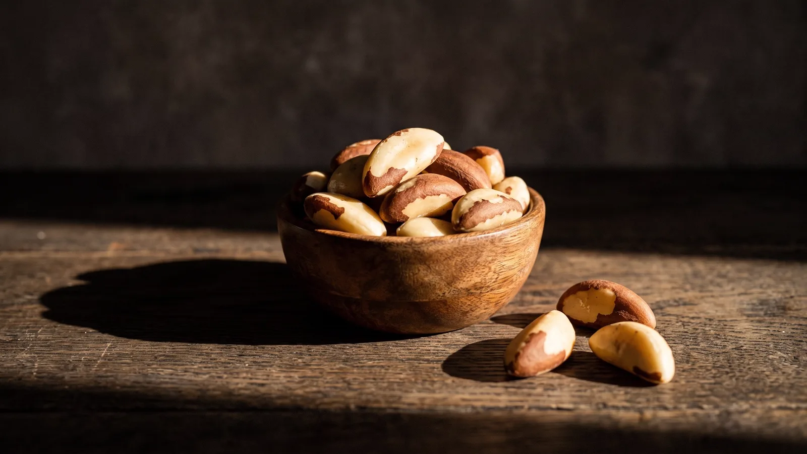 A wooden bowl of shelled Brazil nuts on a dark weathered table in warm light