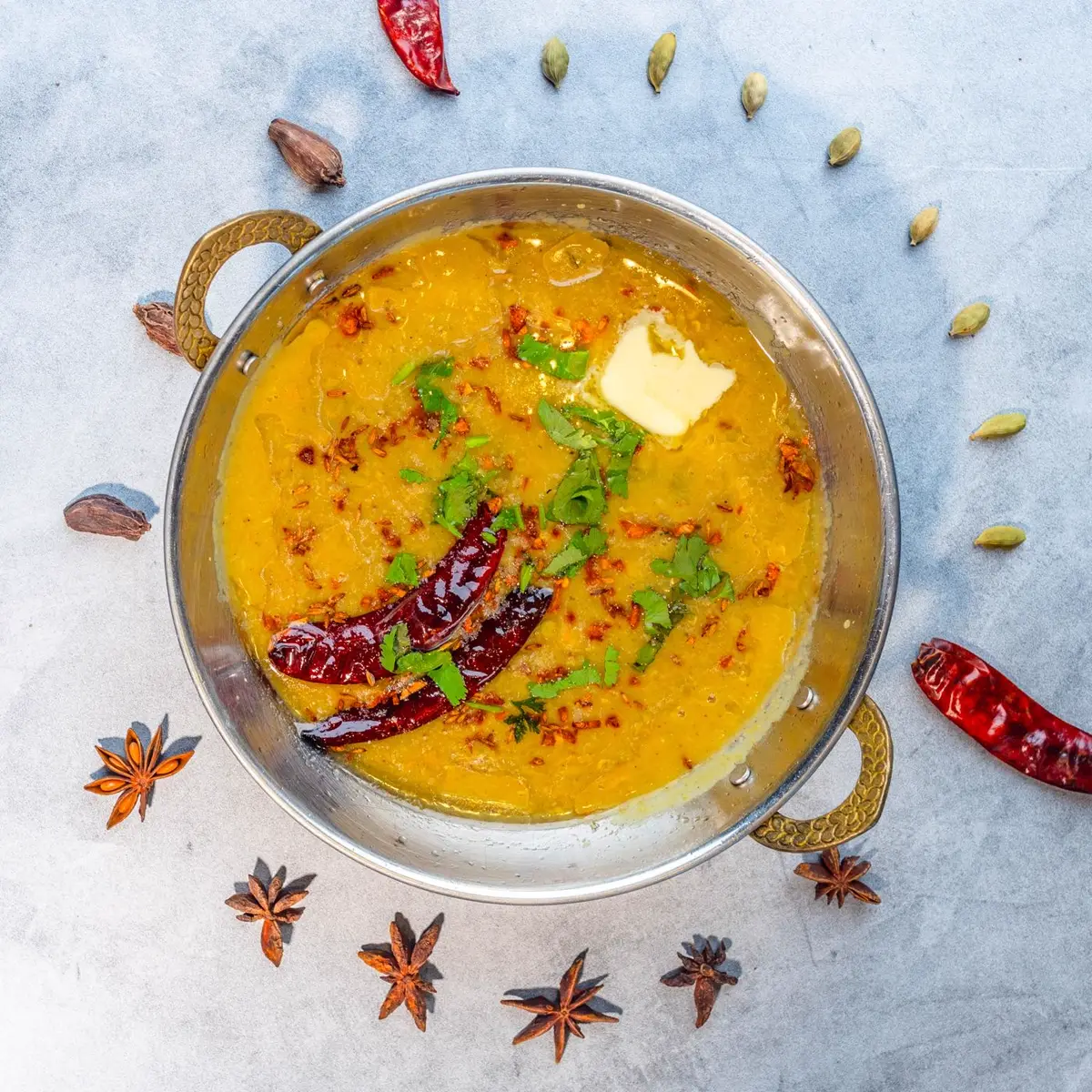 A brass bowl of golden lentil dal with melting fat, dried chilies and fresh cilantro, surrounded by whole spices